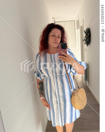 Woman taking mirror selfie in striped linen dress with straw bag. Minimal style, everyday authenticity, and modern self-expression through fashion. Woman taking mirror selfie in striped linen dress with straw bag. Minimal style, everyday authenticity, and modern self-expression through fashion. 133050543