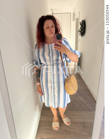 Woman taking mirror selfie in striped linen dress with straw bag. Minimal style, everyday authenticity, and modern self-expression through fashion. Woman taking mirror selfie in striped linen dress with straw bag. Minimal style, everyday authenticity, and modern self-expression through fashion. 133050544