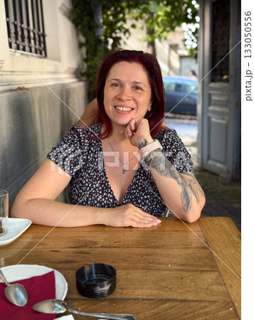 Woman sitting at wooden table in outdoor cafe, relaxed and smiling. Warm sunlight, comfort, and peaceful mood of a slow afternoon. 133050556