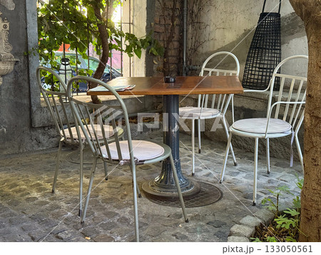 Wooden table and metal chairs under tree shade in outdoor cafe. Peaceful corner, harmony of nature and urban comfort in timeless style. Wooden table and metal chairs under tree shade in outdoor cafe. Peaceful corner, harmony of nature and urban comfort in timeless style. 133050561