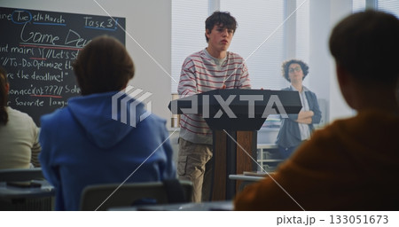 Teenage Boy at Podium Explains Work Peers, Game Dev Topic Visible on Blackboard 133051673