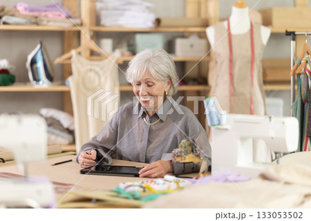 Positive mature female dressmaker drawing in tablet while sitting at table 133053502