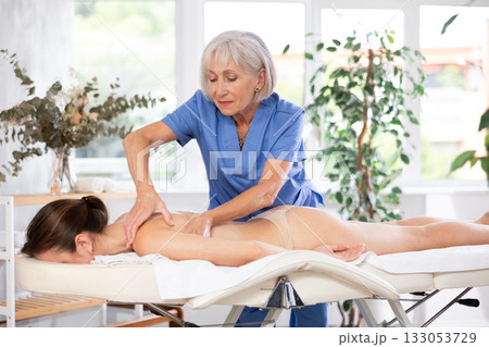 Experienced older professional masseuse doing back massage to young woman Experienced older professional masseuse doing back massage to young woman 133053729