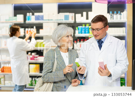 Man apothecary hold package with cream and talks with senior woman client about nuances of care 133053744