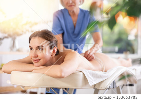 Cropped mature woman actively rubs lower back of young woman during general massage procedure. Cropped mature woman actively rubs lower back of young woman during general massage procedure. 133053785