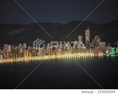 vivid city nightscape, city lights illuminate waterfront against mountain silhouetted backdrop 133058164