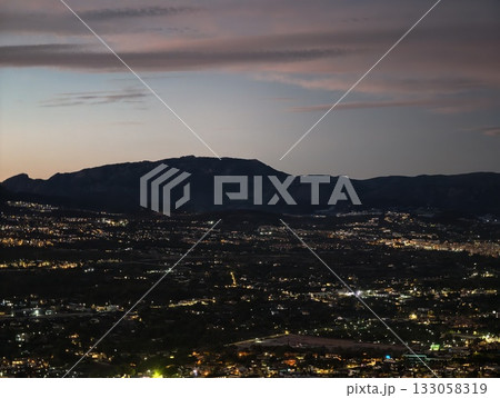 bright cityscape beneath towering mountains, vivid metropolis beyond shadowed mountain ranges 133058319