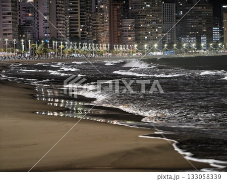 moonlit shore with crashing waves, nighttime seaside view with glowing city lights and rippling surf moonlit shore with crashing waves, nighttime seaside view with glowing city lights and rippling surf 133058339