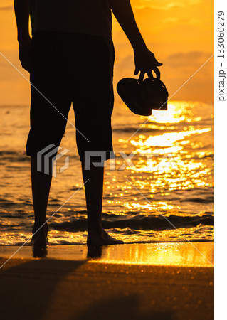 Silhouette of a man standing barefoot on the beach at sunrise, holding sandals in hand as golden sunlight reflects on the waves. 133060279