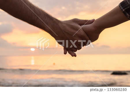 Close-up of two people holding hands against a beautiful sunrise over the sea, symbolizing love, unity, and trust in a serene coastal atmosphere 133060280