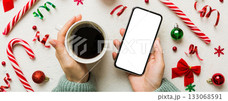 Above view of female hand holding smart phone with hot cup of coffee. Christmas decoration on colored background 133068591