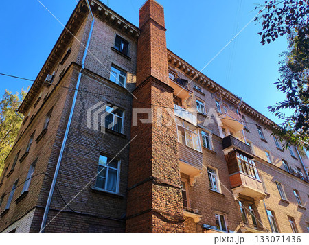 Old multi-story brick building with uneven balconies and a tall red chimney rising along its facade under clear blue sky. The photo conveys the atmosphere of aging Soviet housing, suitable for topics Old multi-story brick building with uneven balconies and a tall red chimney rising along its facade under clear blue sky. The photo conveys the atmosphere of aging Soviet housing, suitable for topics 133071436