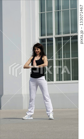A woman in sportswear stands outdoors in a yoga pose, palms pressed together in a moment of focus. The combination of fitness and modern architecture highlights the balance between movement and 133071467