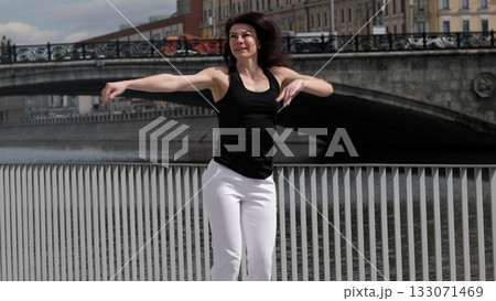 A woman in sportswear gracefully moves near a riverfront, her arms extended in a dynamic motion. The fusion of fitness and urban scenery highlights the energy of an active city lifestyle. A woman in sportswear gracefully moves near a riverfront, her arms extended in a dynamic motion. The fusion of fitness and urban scenery highlights the energy of an active city lifestyle. 133071469