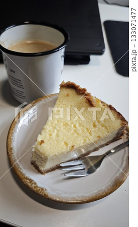 Slice of creamy cheesecake on a plate with a fork next to a cup of coffee. The image conveys comfort, indulgence and a cozy coffee break moment 133071477