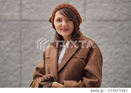 A woman in a brown coat and knitted hat smiles softly outdoors, standing against a textured wall. The image conveys autumn warmth, calm confidence, and natural elegance. 133071481