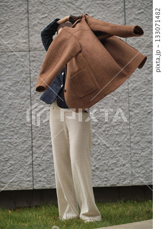 A woman lifts a brown wool coat to put it on, captured in motion against a modern gray wall. The frame reflects autumn warmth, everyday elegance, and timeless fashion in movement. A woman lifts a brown wool coat to put it on, captured in motion against a modern gray wall. The frame reflects autumn warmth, everyday elegance, and timeless fashion in movement. 133071482