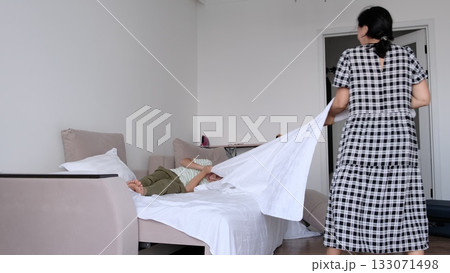 A woman laughs mid motion as she flips a crisp sheet across the bed while her son relaxes, photographed with clean composition. Daily habits shape care, hygiene, and trust in modern family life. 133071498