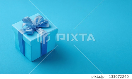A small, sky-blue gift box, tied with a matching ribbon and bow, sits diagonally on a vibrant blue surface.  A shadow adds depth.  Perfect for a celebration or special occasion. 133072240
