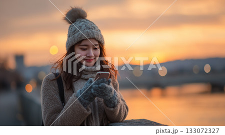 夕暮れに微笑む|冬の川辺でスマートフォンを楽しむ女性 夕暮れに微笑む|冬の川辺でスマートフォンを楽しむ女性 133072327