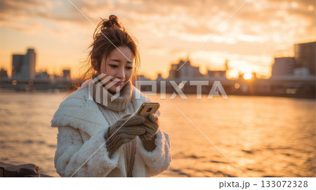夕暮れに微笑む|冬の川辺でスマートフォンを楽しむ女性 夕暮れに微笑む|冬の川辺でスマートフォンを楽しむ女性 133072328