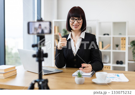 Caucasian woman wearing business attire blogging about professional topics at table with accessories. 133075232