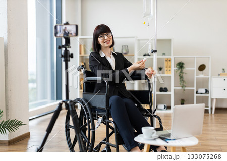 Caucasian woman recording blog with phone while working from wheelchair and receiving IV treatment. Modern office space, smart devices, professional attire, and healthcare equipment Caucasian woman recording blog with phone while working from wheelchair and receiving IV treatment. Modern office space, smart devices, professional attire, and healthcare equipment 133075268