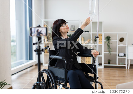 Caucasian woman in wheelchair with intravenous drip recording blog about handling treatment with work. She conveys empowerment and adaptability in modern life, highlighting professional and balance. Caucasian woman in wheelchair with intravenous drip recording blog about handling treatment with work. She conveys empowerment and adaptability in modern life, highlighting professional and balance. 133075279