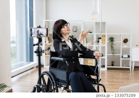 Caucasian woman in wheelchair with intravenous drip recording blog about handling treatment with work. She conveys empowerment and adaptability in modern life, highlighting professional and balance. Caucasian woman in wheelchair with intravenous drip recording blog about handling treatment with work. She conveys empowerment and adaptability in modern life, highlighting professional and balance. 133075289