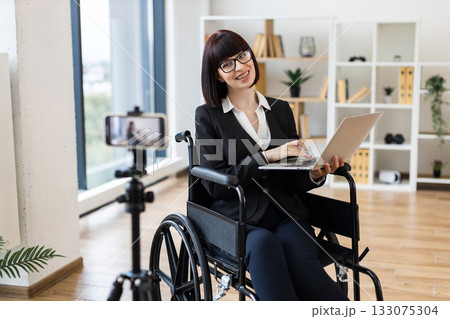Caucasian woman in wheelchair holding laptop blogging about business using camera in office. Caucasian woman in wheelchair holding laptop blogging about business using camera in office. 133075304