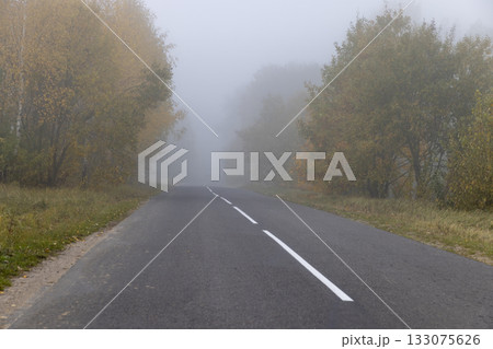 a narrow country road with yellowing trees on the edge of the road in the autumn season, cloudy weather with a gray nondescript sky, fog in the morning due to climate change 133075626