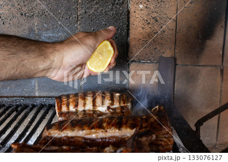 Hand squeezing fresh lemon juice over grilling juicy meat on a hot argentine barbecue, enhancing flavors and creating a healthy, appetizing meal outdoors Hand squeezing fresh lemon juice over grilling juicy meat on a hot argentine barbecue, enhancing flavors and creating a healthy, appetizing meal outdoors 133076127