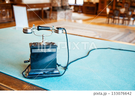 An old antique vintage telephone placed on a wooden table. The classic rotary phone with elegant design and metal details reflects historical charm and retro aesthetics. Perfect image for illustrating An old antique vintage telephone placed on a wooden table. The classic rotary phone with elegant design and metal details reflects historical charm and retro aesthetics. Perfect image for illustrating 133076642