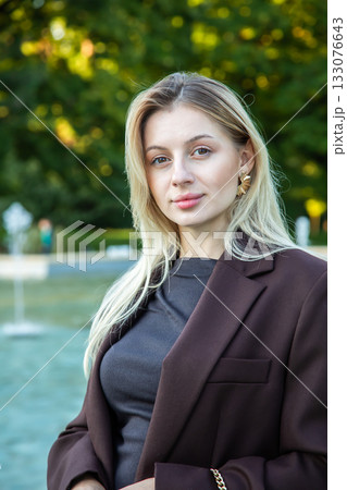 Young attractive blonde businesswoman standing near a city fountain in autumn. Stylish woman in a burgundy blazer, confident and elegant, enjoying a sunny fall day outdoors. Perfect for lifestyle Young attractive blonde businesswoman standing near a city fountain in autumn. Stylish woman in a burgundy blazer, confident and elegant, enjoying a sunny fall day outdoors. Perfect for lifestyle 133076643