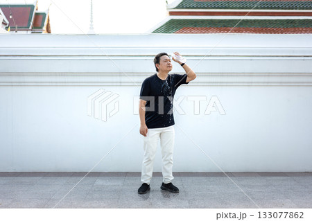 Thai man pours drinking water over his face to cool down under the intense Bangkok heat. Standing against a temple wall, he refreshes himself in a moment of relief from the scorching day. Thai man pours drinking water over his face to cool down under the intense Bangkok heat. Standing against a temple wall, he refreshes himself in a moment of relief from the scorching day. 133077862