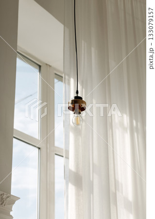 Modern hanging light fixture with soft natural light through sheer curtains and large window 133079157