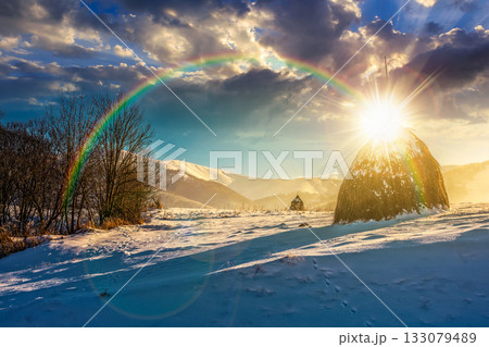 rural scenery with haystack in winter at sunset. countryside landscape with snow covered hill in mountains in evening light. low angle view with snowy peak in the background. storytelling image under 133079489