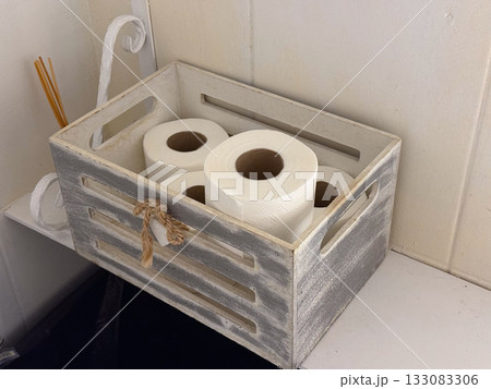 Wooden crate with toilet paper rolls on white shelf. Simple bathroom storage, everyday organization, and minimal hygiene detail. 133083306