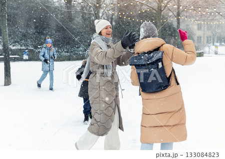 Group young adult smiling women family wear long quilted coat white hat enjoy play snowball snowy day outdoors friends. Snowflakes fall playfully in front stairs decorated with glowing winter lights 133084823