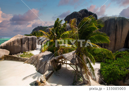 Granite boulders frame a narrow sandy path to turquoise water at left. Coconut palms and a thatched shelter sit on powder sand in warm late afternoon light. 133085003