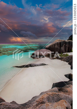 Crescent white sand and weathered granite at La Digue, Seychelles, face a teal lagoon. Pink and amber storm clouds move at dusk. Wide angle long exposure feel. 133085013