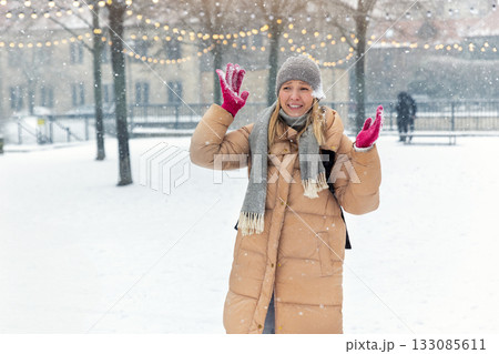 Portrait young adult smiling woman wear long quilted coat white hat enjoy play snowball snowy day outdoors. Snowflakes fall she moves playfully in front of stairs decorated with glowing winter lights Portrait young adult smiling woman wear long quilted coat white hat enjoy play snowball snowy day outdoors. Snowflakes fall she moves playfully in front of stairs decorated with glowing winter lights 133085611
