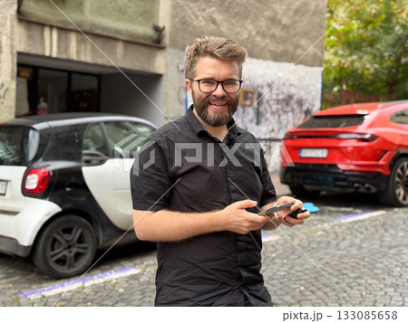 Man using smartphone on city street near parked cars. Technology, urban lifestyle, and everyday communication in modern environment. 133085658