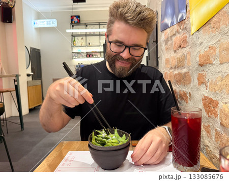 Man eating edamame with chopsticks in cafe. Modern dining culture, healthy lifestyle, and leisure moment in urban restaurant. 133085676