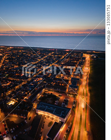 Aerial dusk view of coastal town warm streetlights trace diagonal road past dense buildings and modern complex, ocean horizon glows orange pink beneath deep blue sky with scattered clouds. Aerial dusk view of coastal town warm streetlights trace diagonal road past dense buildings and modern complex, ocean horizon glows orange pink beneath deep blue sky with scattered clouds. 133085751