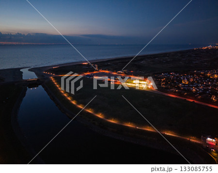 Aerial twilight view of coastal town streetlights trace canal and curved shoreline, glowing path leads to illuminated buildings, serene water and distant horizon blend natural calm with urban order. Aerial twilight view of coastal town streetlights trace canal and curved shoreline, glowing path leads to illuminated buildings, serene water and distant horizon blend natural calm with urban order. 133085755