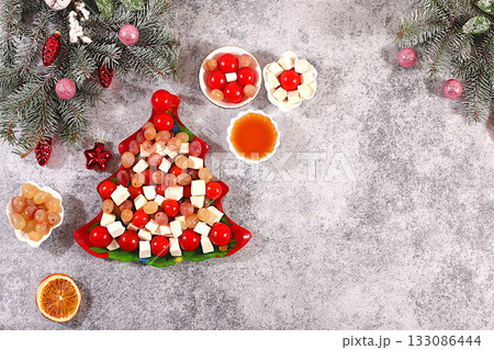 Christmas and New Year's dishes, traditional holiday salad in the shape of a Christmas tree with feta cheese, tomatoes, grapes, olives and honey with fir branches and decorations, dish decoration idea Christmas and New Year's dishes, traditional holiday salad in the shape of a Christmas tree with feta cheese, tomatoes, grapes, olives and honey with fir branches and decorations, dish decoration idea 133086444