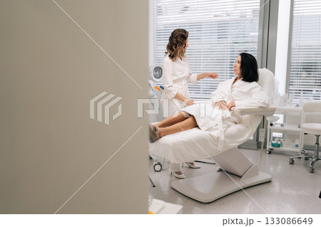 Remote view of woman client in bathrobe attentively listening to cosmetologist during in-clinic consultation about personalized aesthetic treatments, skin care plans and wellness options. Remote view of woman client in bathrobe attentively listening to cosmetologist during in-clinic consultation about personalized aesthetic treatments, skin care plans and wellness options. 133086649