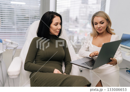 Cosmetologist presenting information on laptop to female client, analyzing skincare needs and discussing potential treatments in a modern aesthetic clinic. Concept of beauty treatment. 133086653