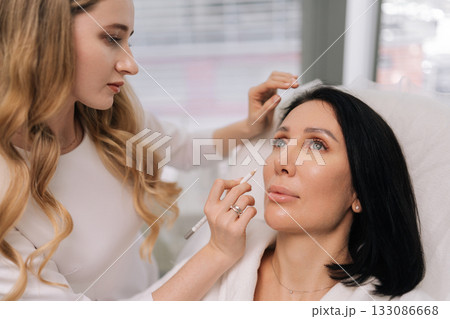 Close-up face of middle-aged woman receiving facial aesthetic procedure featuring cosmetologist marking face with pencil before beauty treatment, preparing for rejuvenation in modern beauty clinic. Close-up face of middle-aged woman receiving facial aesthetic procedure featuring cosmetologist marking face with pencil before beauty treatment, preparing for rejuvenation in modern beauty clinic. 133086668